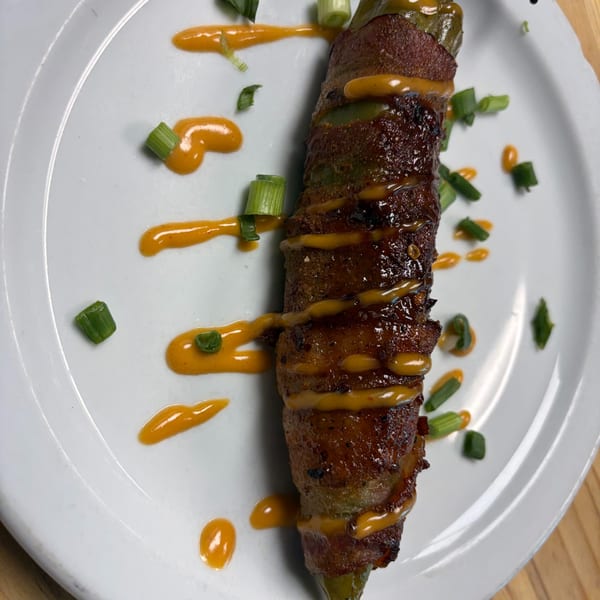 Bacon-wrapped jalapeño popper drizzled with orange sauce and garnished with green onions on a white plate.
