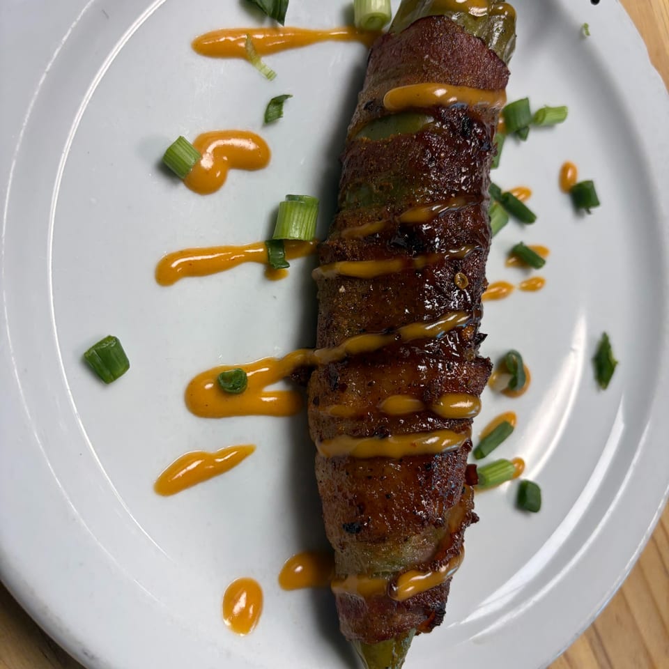 Bacon-wrapped jalapeño popper drizzled with orange sauce and garnished with green onions on a white plate.