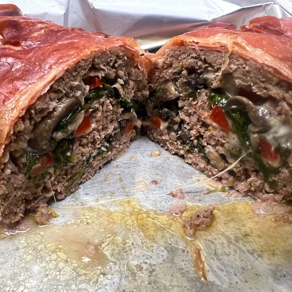 A bacon-wrapped meatloaf, cut in half, revealing a savory filling of spinach, mushrooms, and red peppers.