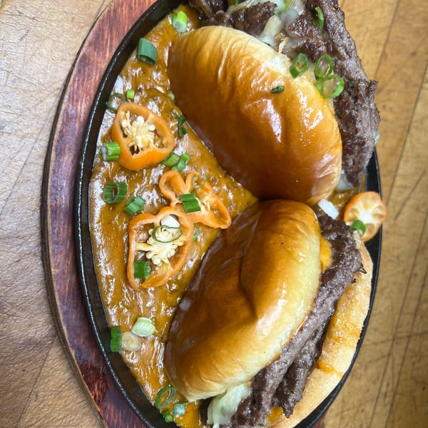Two cheesesteak sliders in a skillet with melted cheese sauce, sliced peppers, and green onions.