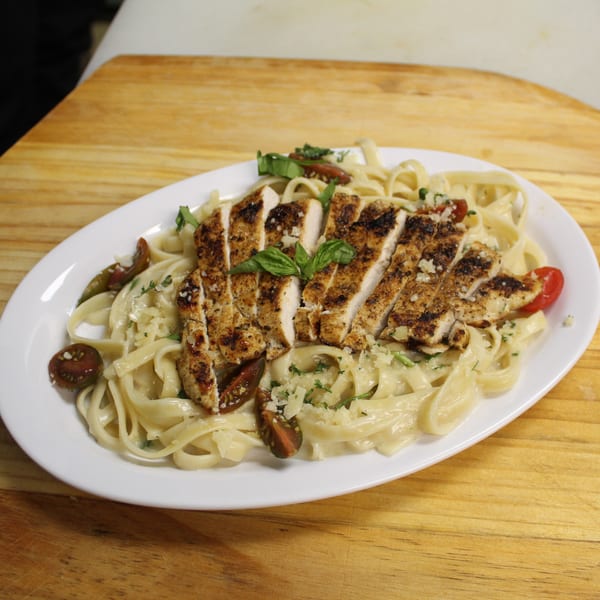 Blackened chicken fettuccine alfredo with cherry tomatoes, basil, and parmesan cheese on a white plate.
